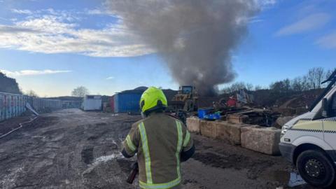 The back of a firefighter in protective gear in what looks like a building yard. There is black smoke rising into the air. 