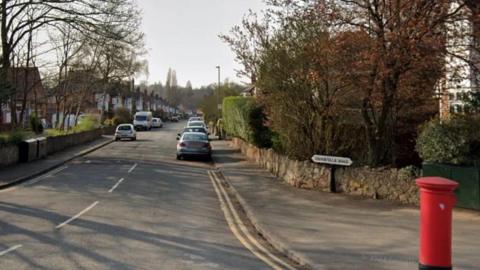 A street view image of Brookvale Road. The street is lined with trees, homes and has cars parked on either side of the road. A red post box is in the foreground of the image. 