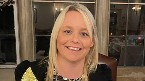 April Jeffers smiles facing the camera as she sits in a room with stone walls and large windows. She has straight, blonde hair below her shoulders, wears a black dress or top and holds a golden trophy from an awards ceremony.