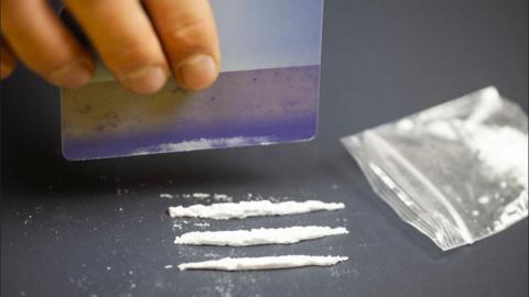 A hand holds a plastic credit card above three lines of white powder on a table top next to a small empty plastic bag