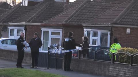 Five police officers stand guard in front of a property which has been cordoned off. The house is fronted by a metal rail and brick wall which has blue and white police tape attached to it. A row of other houses are visible in the background.
