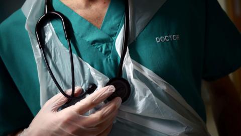 A stock image of a doctor wearing a plastic apron over their scrubs and holding a stethoscope.