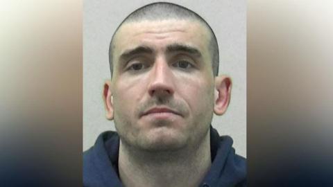 Mugshot of Wylie. He has shaved dark hair and very faint dark stubble and is wearing a navy hoodie.
