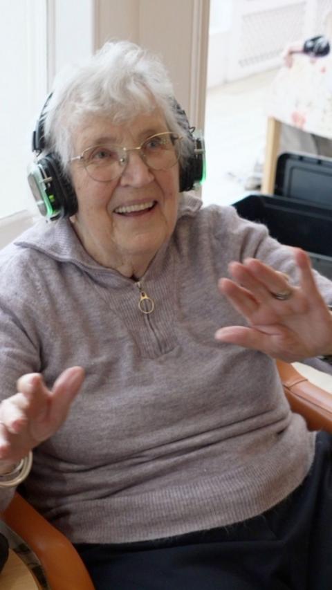 An elderly lady sitting in a chair wearing a grey jumper smiles as she takes part in a silent disco in a care home. She is wearing headphones and is waving her arms