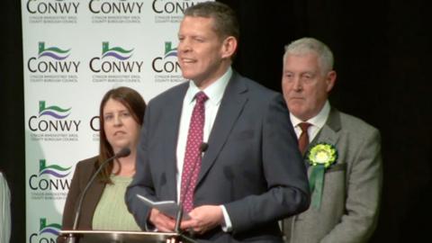Rhun ap Iorwerth, Plaid Cymru leader talks into a crowd with two people behind him. 
