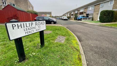 A white street sign with black writing saying "Philip Road, leading to Percy Way". The sign stands on a grass verge. There are residential buildings and parked cars in the background.