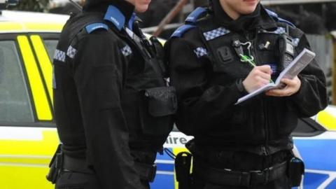 Two police officers wearing black standing in front of a police car holding note pads