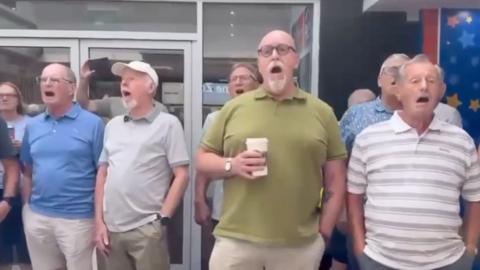 Multiple men are seen singing at a service station. From right to left one man is wearing glasses, a blue t shirt and has shoulder length hair, the next man has glasses, he is wearing a blue polo shirt and cream trousers. The next man has a cream baseball cap, a grey polo shirt and green trousers. The next is a man in the background, he is wearing glasses and a white shirt. The man nearest to the camera is wearing an olive green polo shirt and cream trousers, then the last man furthest to the left is wearing a white and grey striped polo shirt and gray trousers. The rest of the men in the background are barely in view but there is one man with a light blue polo shirt with a spotted pattern who is predominantly blocked by another.
