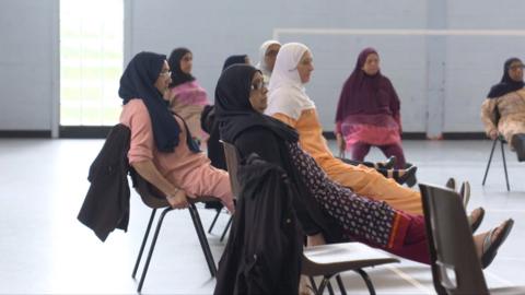 South Asian widows exercising together in Blackburn. Image shows women doing chair yoga in a community centre.