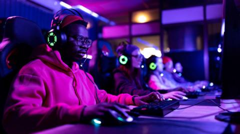 A man is playing a video game on a computer, there are other gamers, wearing the same green illuminated headphones, out of focus along the desk to his left.