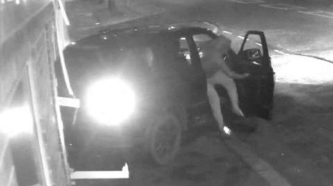 Black and white CCTV still showing a black Range Rover reversed into a building and a man with a hood up jumping out of the driver's seat