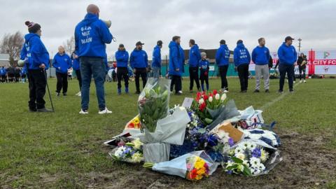 Lots of bunches of flowers laying on a football pitch whilst community members behind are wearing blue hoodies that say 'R.I.P Max and Mason'