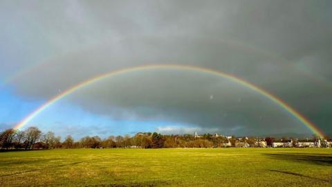 A full rainbow sits right across an open field with dark grey clouds and a small patch of blue sky in the left