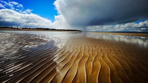 Vertical ridge lines run across brown and orange sand with a huge white cloud above on the right and white clouds and blue sky on the left