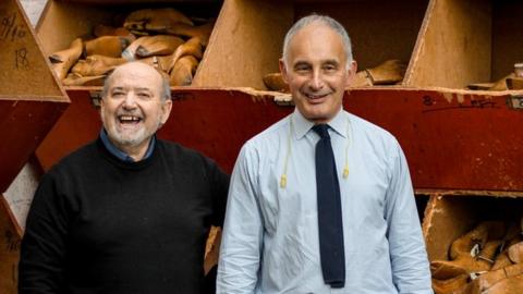 Two men, one wearing a tie and the other a blue sweater, stand in front of containers of part-built shoes