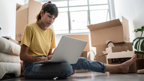 Person inside using laptop while sitting on floor with cardboard boxes in living room after moving house