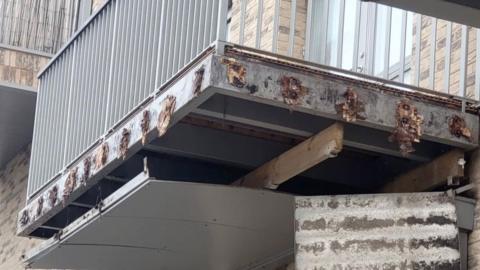 A damaged balcony on the Weavers estate, with exposed structural supports and metal sheeting hanging down