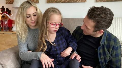 Minnie, with long blonde hair and red glasses, sits between her mother Charlotte and father Jesse