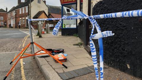 Police tape and defibrillator kit on pavement in Bridge Street, Loddon