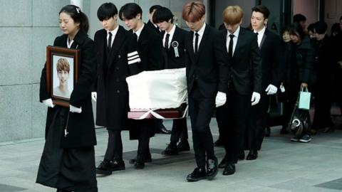 K-pop singers carry the coffin of Jonghyun while his sister walks in front, carrying his photo