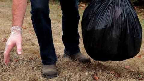 A man wearing a glove reaches to pick up litter as he holds a black bin bag