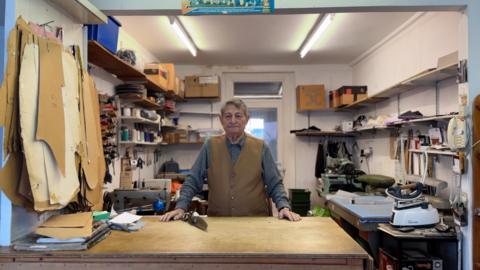 A man in a waistcoat and shirt stands behind the counter in a small tailoring shop