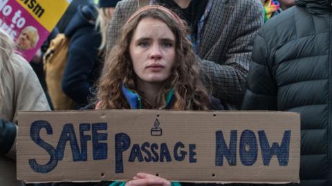 A woman looks sad holding a poster saying safe passage now.