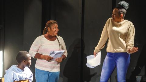 Generic image of a group of actors rehearsing on stage. A young black boy wearing a pale blue hoody sits on a chair. A young black women with long braids stands next to him holding a script and another woman stands beside her also holding a script and posing.