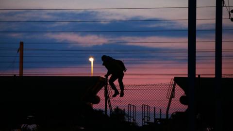 Migrant climbing over a fence at the Eurotunnel terminal at Coquelles