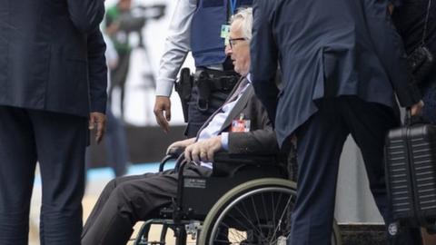 Jean-Claude Juncker is seen seated in a wheelchair as he is brought in through a side entrance of the building