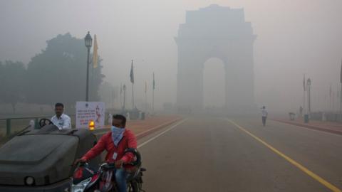 A man covers his face with a scarf as he rides in front of the landmark India Gate, enveloped by smoke and smog, on the morning following Diwali festival in New Delhi