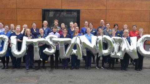 Staff stand outside holding balloons that read 'Outstanding'