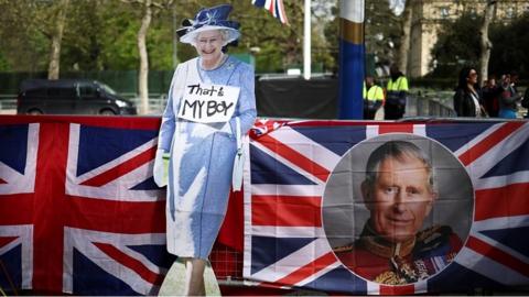 A cardboard cut out of Queen Elizabeth II next to a flag with King Charles III face on, along the coronation route.