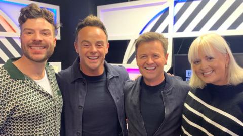 Radio 1 presenters Jordan North and Katie Thistleton stand either side of TV presenters Ant McPartlin and Declan Donnelly. Jordan (left) is a 33-year-old white man with short curly brown hair and wears a patterned green shirt. Ant is a 48-year-old white man with brown hair cut short at the sides - he smiles at the camera and wears an unbuttoned navy shirt over a navy T-shirt. Standing next to Ant is Dec, 48, who is a bit shorter and has slightly lighter brown hair and wears a grey bomber jacket over a blue top. Katie stands on the right - she is a 34-year-old white woman with bleached blonde hair and wears a blue and white striped top. The group are pictured inside a Radio 1 studio.