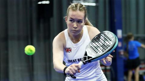 Tia Norton, a woman wearing a white sports vest jersey with Great Britain written on it. She is holding a black padel bat which has a green outer rim.