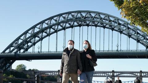 Couple wearing masks walking along Newcastle Quayside with Tyne Bridge in the background