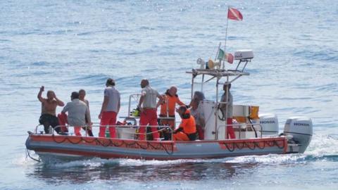 Rescuers survey the site where a luxury yacht sank off the coast of Sicily in an orange boat and dressed in reflective pants.