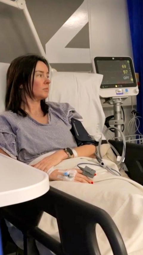 A female patient with shoulder length brown hair sits in a hospital bed wearing a grey t shirt. She has a blood pressure monitor wrapped around her arm and there is a screen in the background.