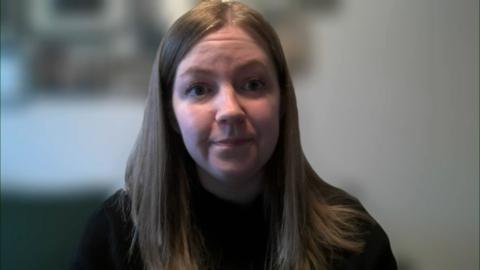 Gillian Mackay sits looking at the camera on a Zoom call, the background is blurred, she has long brown hair and wears a black jumper