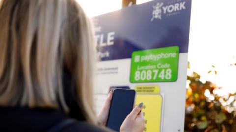 A head and shoulders shot from behind of a woman with light coloured hair. She is holding a mobile phone up in front of a pay and display parking sign.