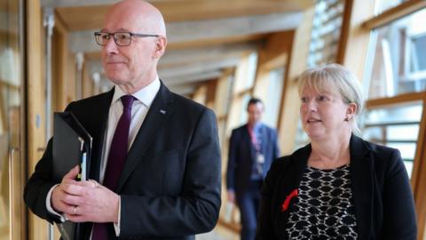 John Swinney, who is bald with glasses, and Shona Robison, who has blonde hair tied back, side by side in the Scottish Parliament. They are both wearing dark jackets, with Swinney carrying a black folder under his arm. There are in a wood-panelled corridor with large windows. 