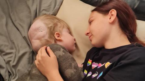 Freya with her brother Harry lying down having a cuddle. Freya has a t shirt with hearts on it and Harry is wearing a brown top with dinosaurs on it.