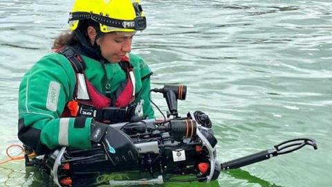 Christel Oweis holds the drone in the water. She wears the HART rescue uniform and a yellow hard hat.