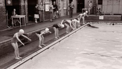 A group of children wearing swimming caps and swimsuits stand at the edge of an outdoor pool, bending forward in starting positions. The poolside is made of brick, and the water is calm. Several adults stand in the background near a building with open doorways, watching the scene. A person sits at a small table to the left with papers in front of them. Signs showing pool depth are fixed to the wall behind. The photo is in black and white.