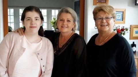 The three woman are standing in a home, smiling at the camera. Lisa has her arm around Chloe, who is wearing a pink top and cardigan. Lisa and Jeannette are in a black top and both wearing a necklace. 