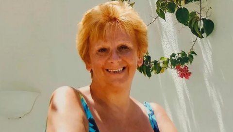 Woman with auburn hair smiling in a sunny setting with a white wall behind her.
