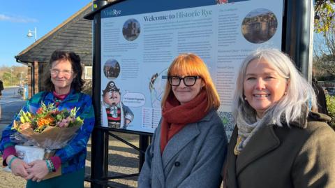 The woman with flowers is Isabel Ryan, daughter of John Ryan. The woman with orange scarf is Sarah Broadbent, Vice Chair of Rye Chamber, with Chair of Rye Chamber, Jane Brook on the right. Behind the women is a sign stating 'Welcome to Historic Rye'. 
