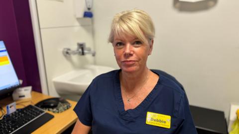 Emergency nurse practitioner, Debbie Chamberlain, looks straight at the camera. She is wearing a dark blue outfit and has a yellow badge which says 'Debbie.' A computer and a sink can be seen in the background.