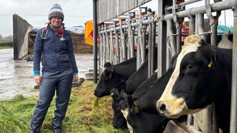 A vet stands in a farmyard. She is dressed in a navy top and waterproof trousers with a colourful bobble hat. She has a stethoscope around her neck and a wide smile. Five cow heads poke through the bars of their shed to reach some grass on the ground. The dairy cow biggest in frame is side-eying the camera.
