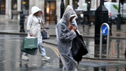 People wrapped in rain macs walk down a street in London carrying bags earlier in the summer.